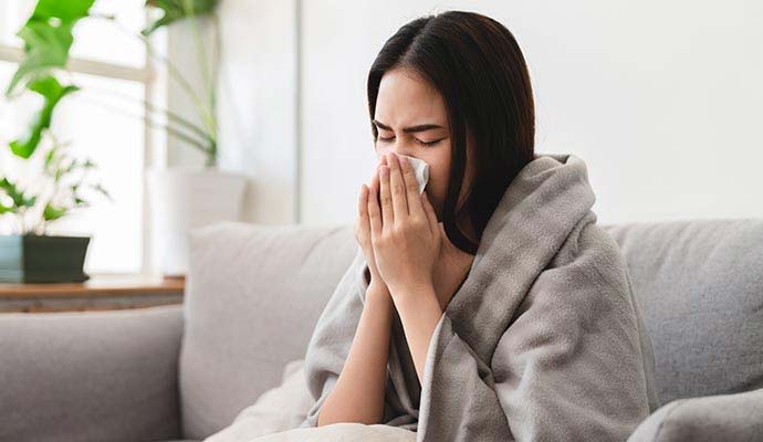 A young woman sitting on a fabric sofa, sneezing into a tissue