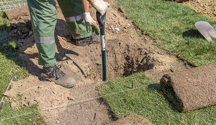 Person digging in a yard with rolls of fresh sod ready for installation as part of a landscaping project